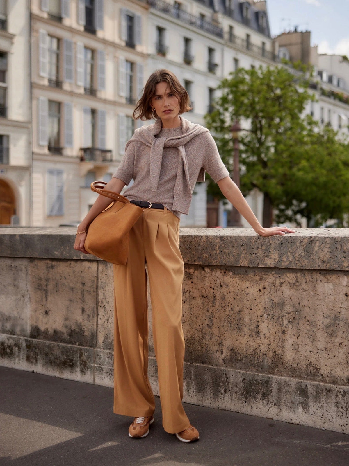 Woman in camel wide-leg trousers and a taupe blazer leaning against a stone bridge railing with Parisian buildings in the background