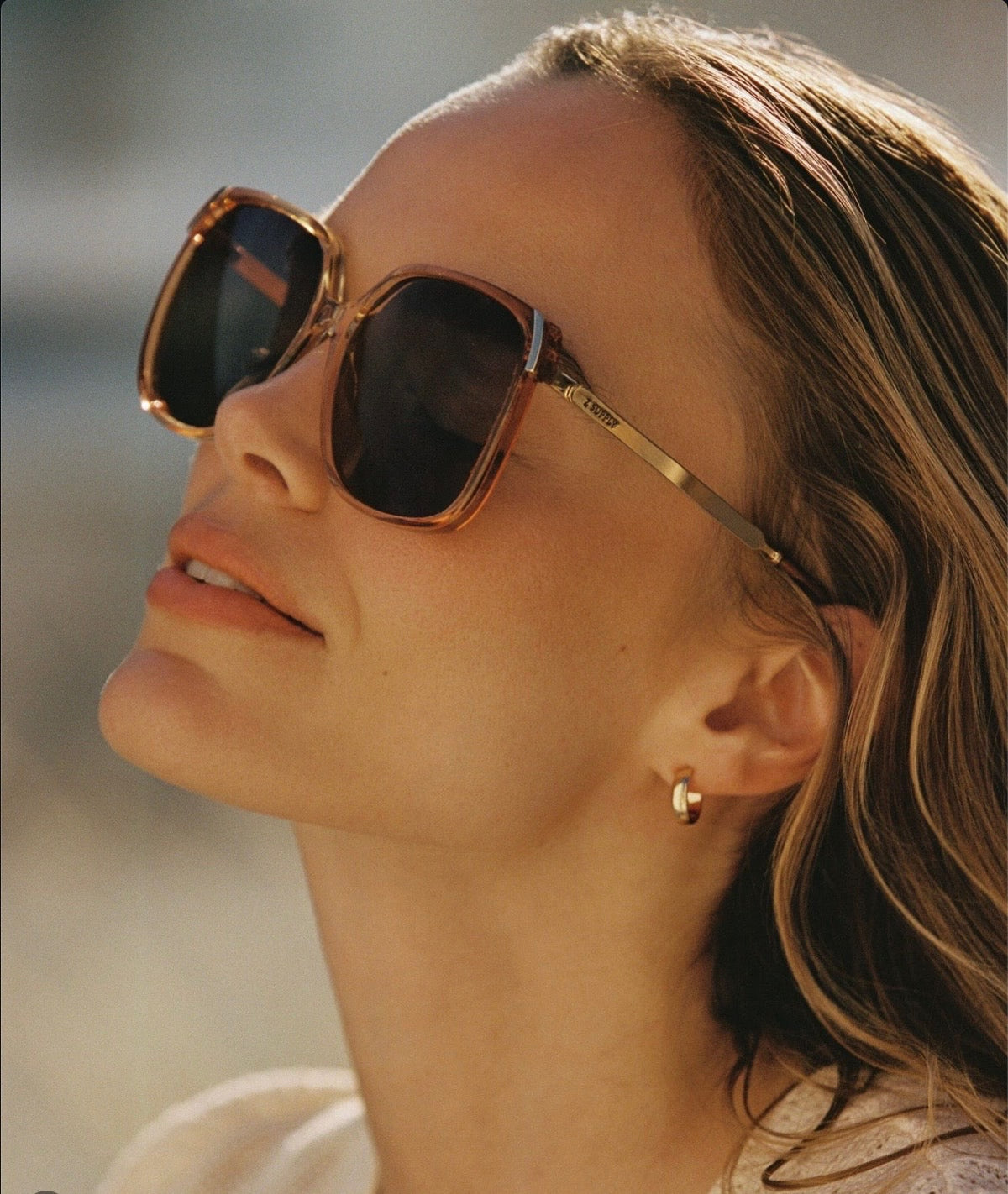 Woman wearing oversized tortoiseshell square sunglasses with gold earrings, looking up in warm sunlight