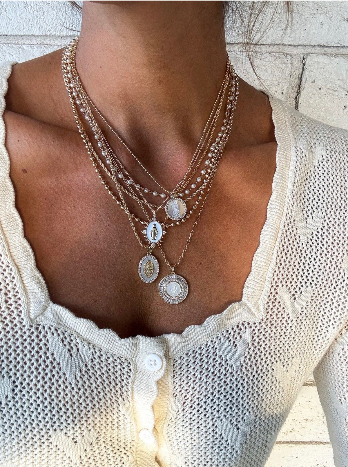 Close-up of layered gold and silver necklaces with pearl and coin pendants worn over a cream knit top