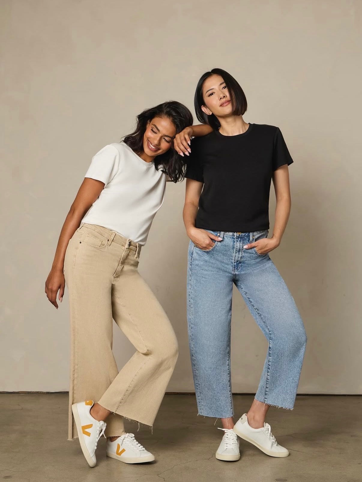 Two women posing together wearing casual jeans — one in tan wide-leg jeans with a white tee, one in blue straight-leg jeans with a black tee and white sneakers