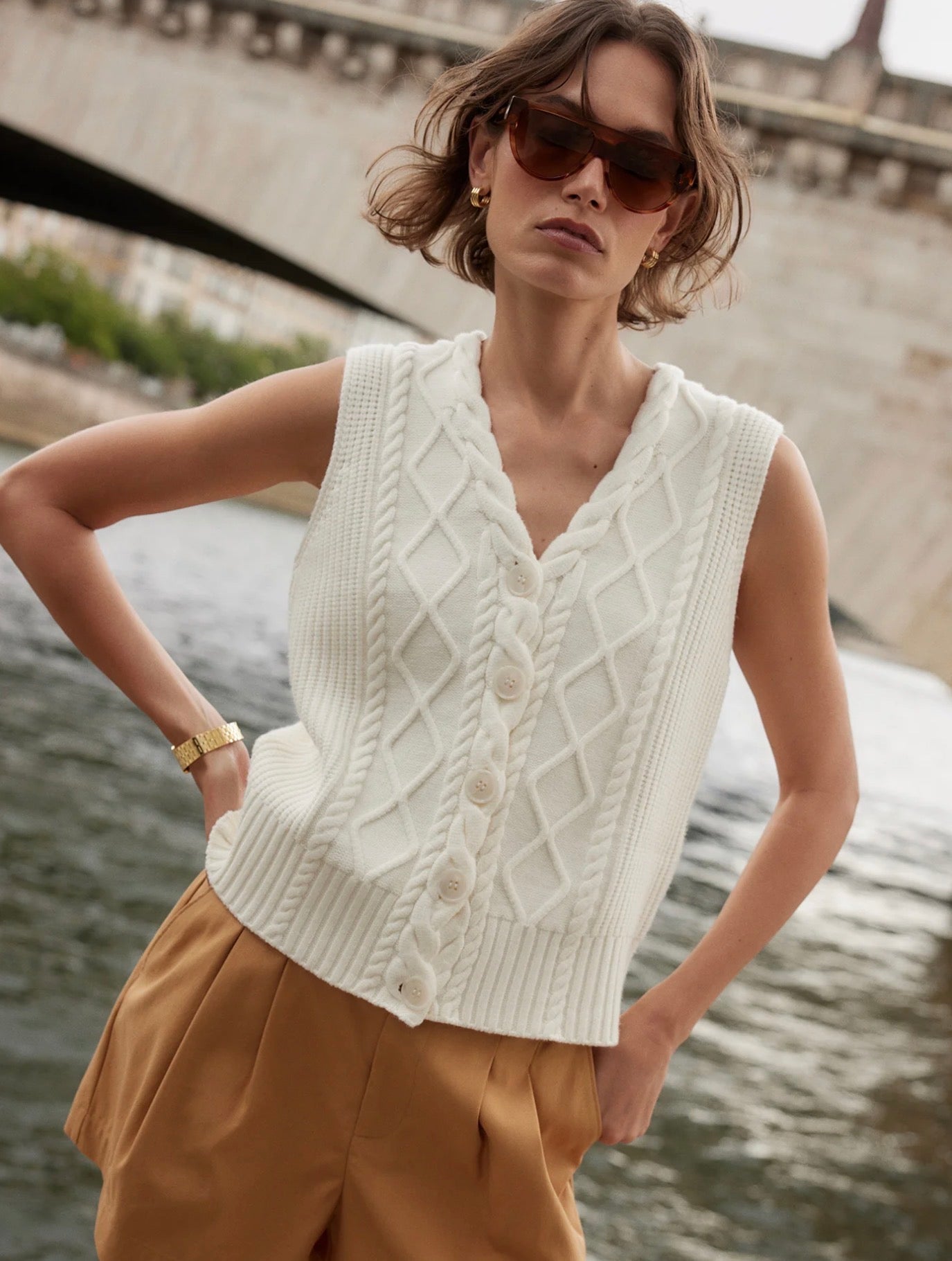 Woman wearing a white knitted vest and sunglasses by a river with a bridge in the background